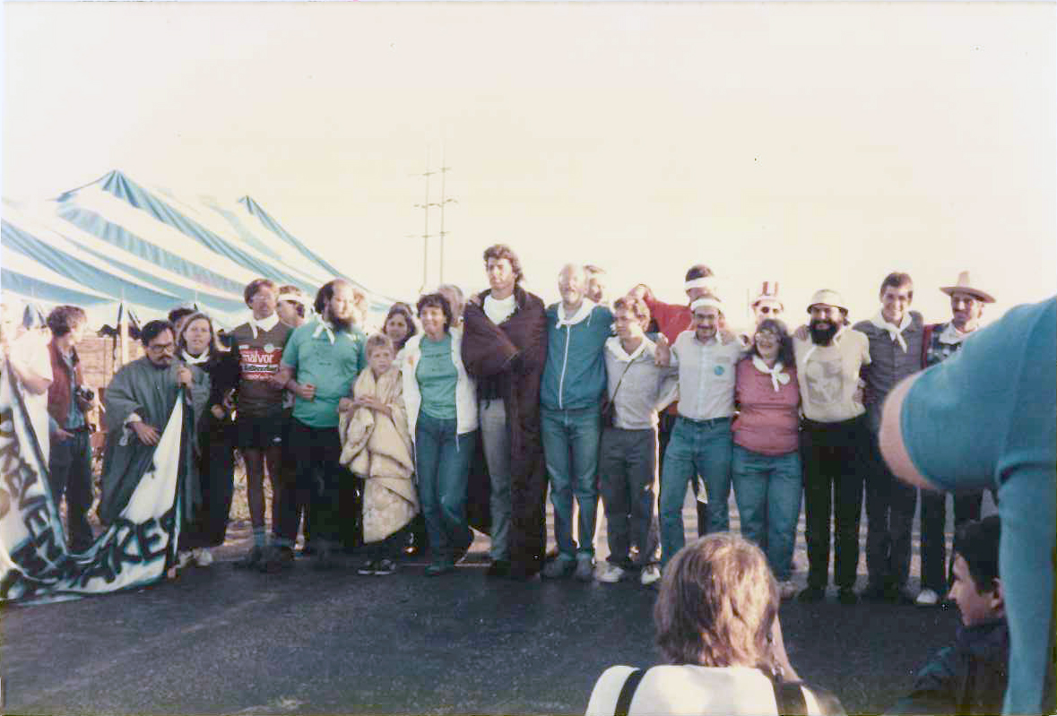 blockading the road to the Pantex gate during an August Peace Camp, some stayed to be arrested. Bob Henschen is in aqua jacket, just right of the tallest person in the line who has a brown blanket around his shoulders, and Mavis Belisle in pink t-shirt fourth from right end
